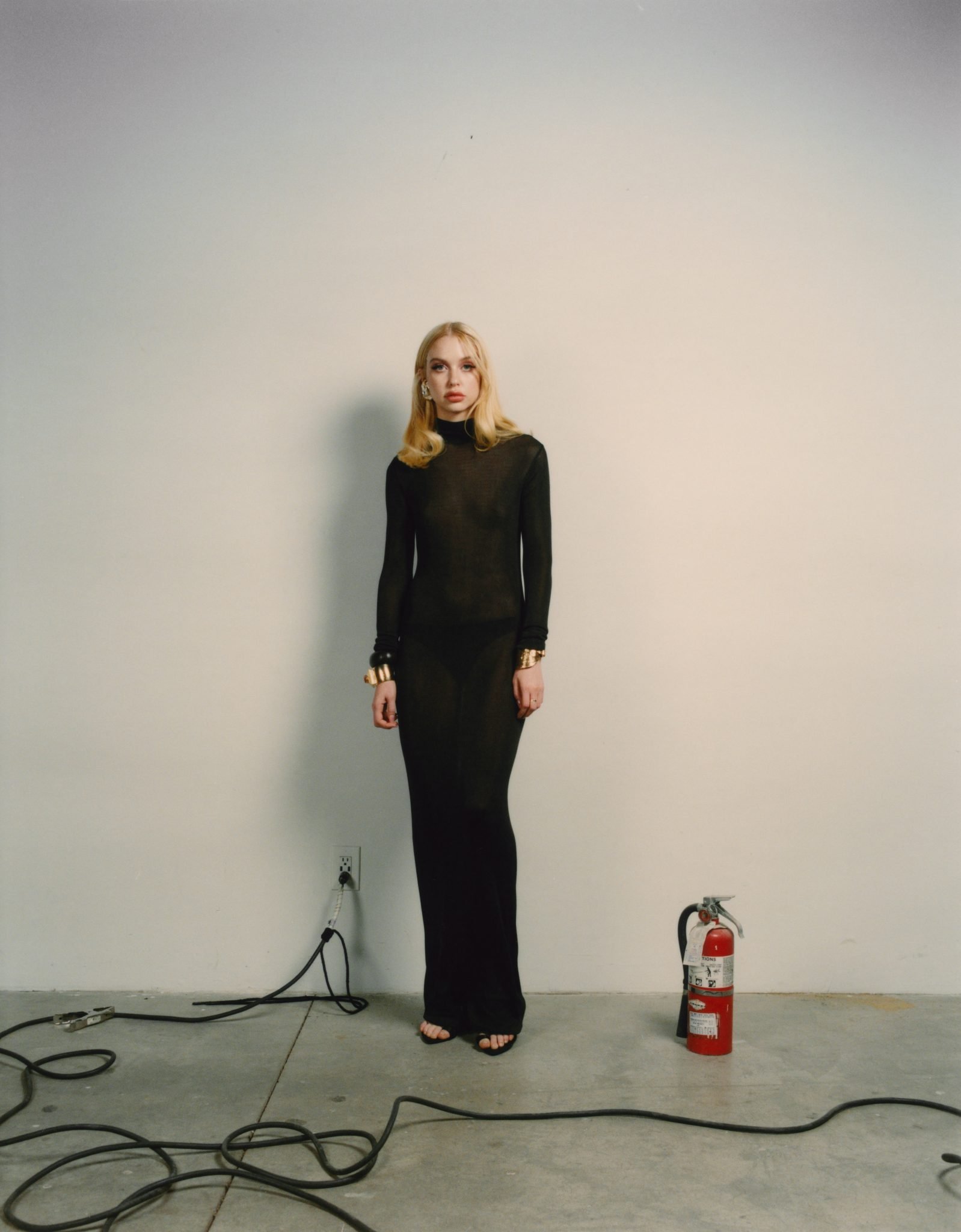 Full-length portrait of a blonde woman in a sheer black dress with gold bracelets, posing in front of a white wall in a photo studio.
