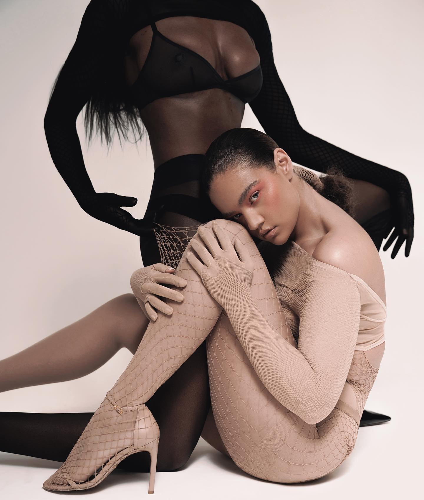 Two women posing in a photo studio wearing lingerie, fishnet stockings, and gloves against a white backdrop, showcasing fashion and beauty.