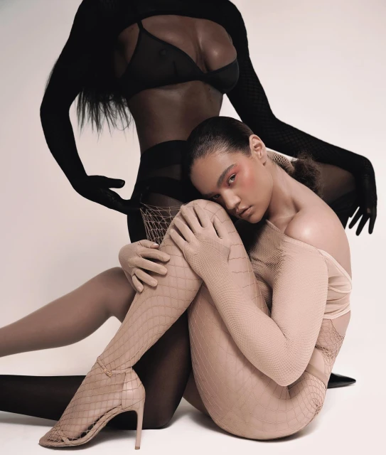 Two women posing in a photo studio wearing lingerie, fishnet stockings, and gloves against a white backdrop, showcasing fashion and beauty.