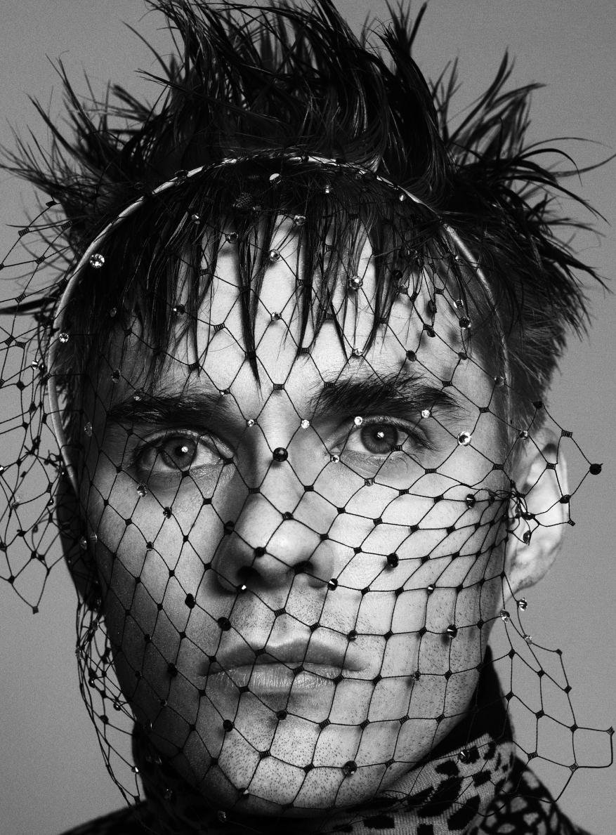 Monochrome close-up of a person wearing a beaded veil and dark patterned shirt, with spiky hair and light eyes in a studio setting.