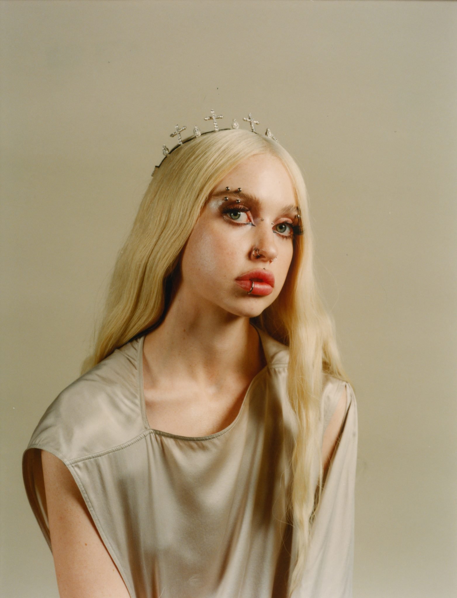 Young woman with blonde hair, wearing a cross crown and a gray sleeveless dress, looks at the camera against a neutral background.