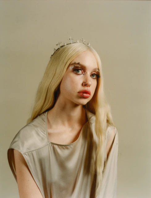 Young woman with blonde hair, wearing a cross crown and a gray sleeveless dress, looks at the camera against a neutral background.