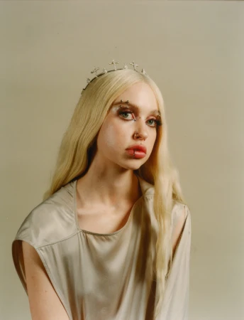 Young woman with blonde hair, wearing a cross crown and a gray sleeveless dress, looks at the camera against a neutral background.