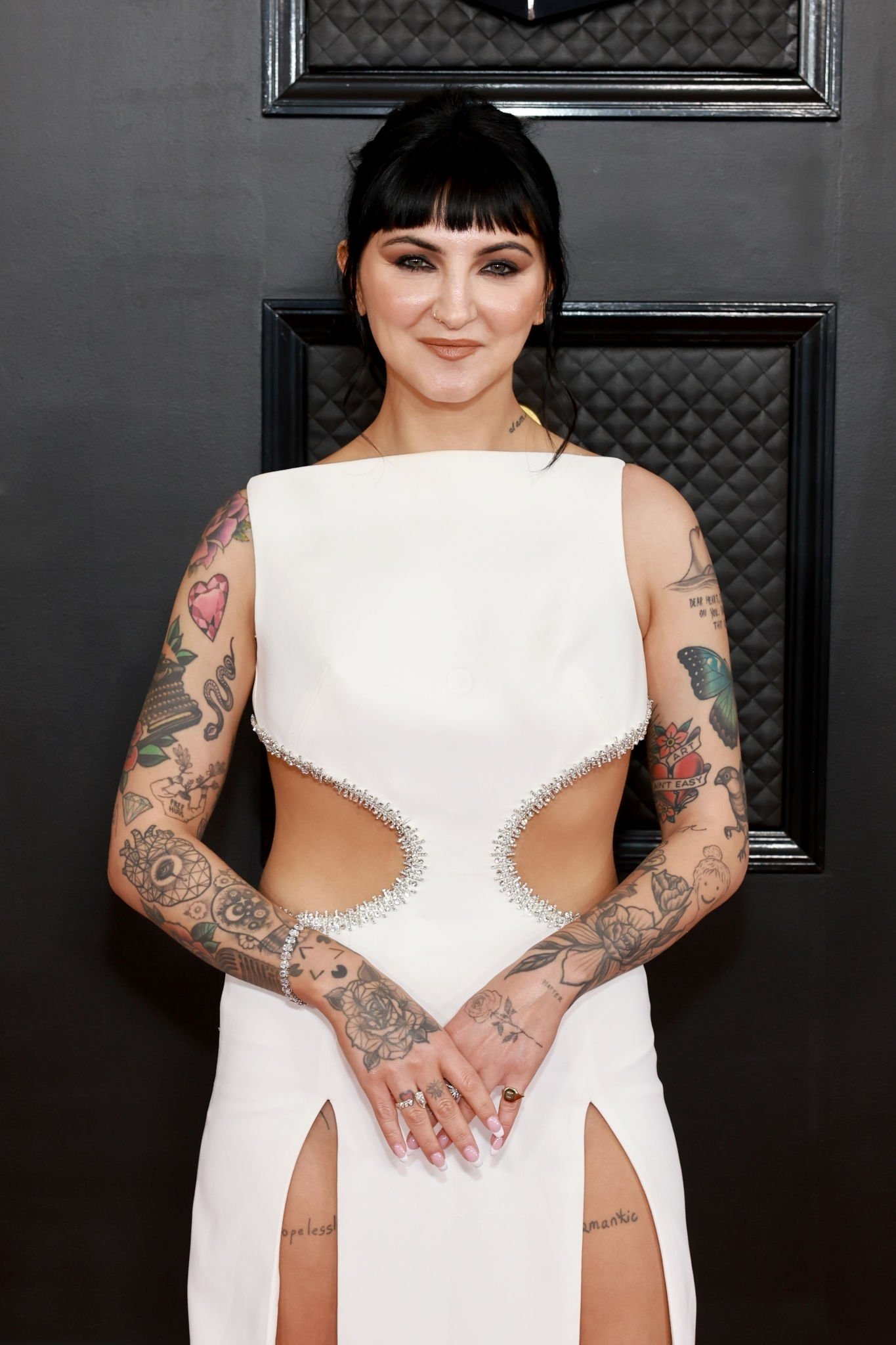 Woman with dark hair and heavy tattoos wearing a white cutout dress standing against a dark quilted backdrop