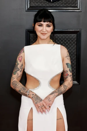 Woman with dark hair and heavy tattoos wearing a white cutout dress standing against a dark quilted backdrop