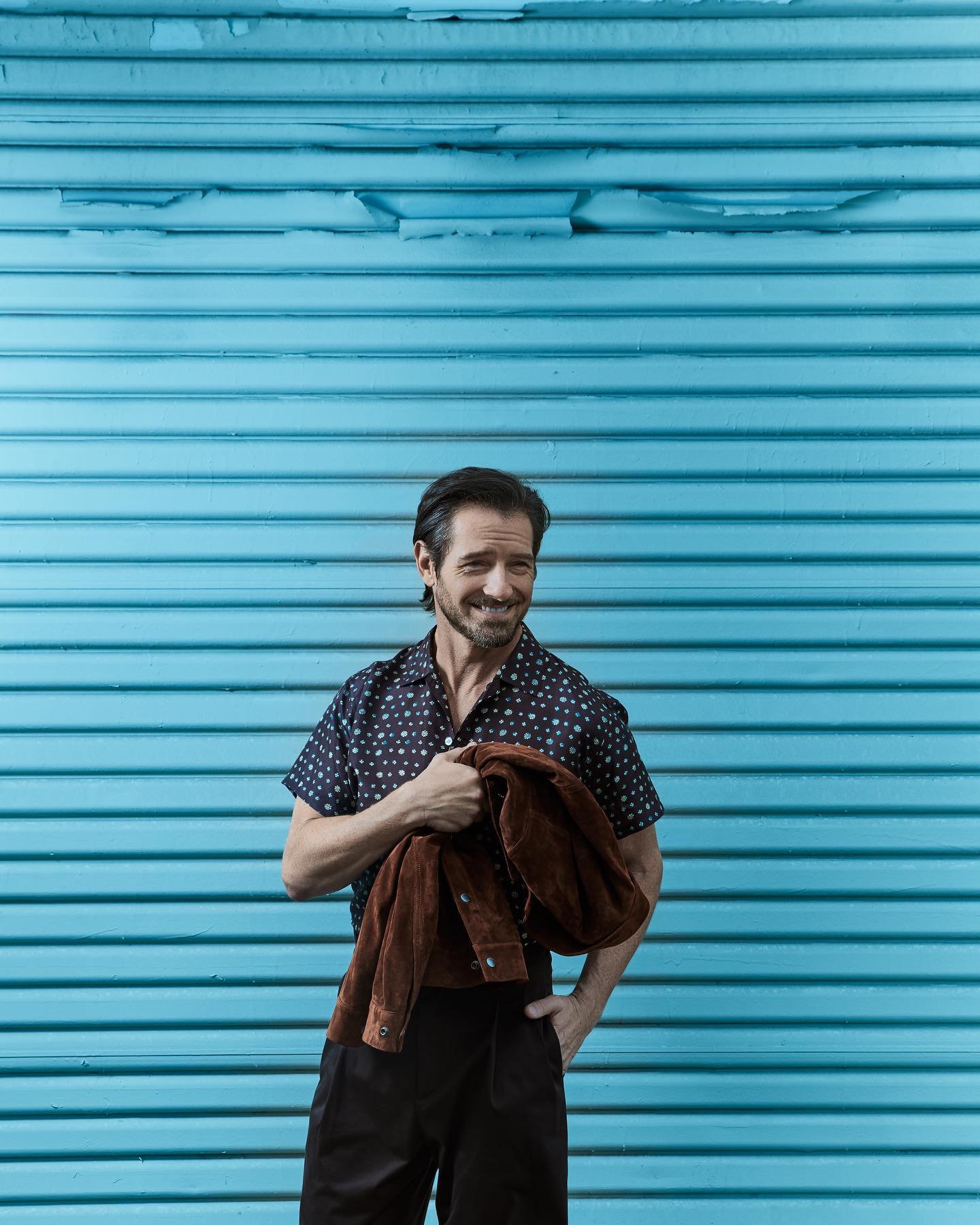 Adult man poses in front of a turquoise blue horizontally striped wall, wearing a patterned shirt and dark slacks while holding a brown jacket.