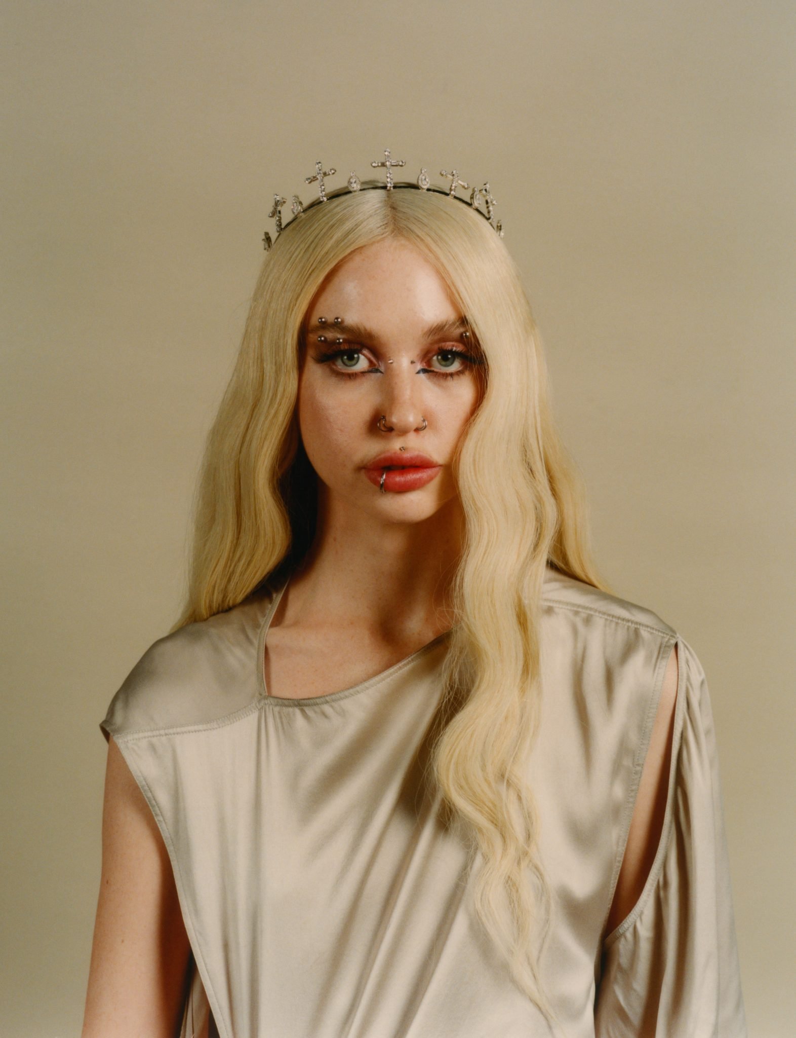 Portrait of a young adult woman with wavy blonde hair wearing a silver crown, a silver dress, and face piercings against a neutral backdrop.