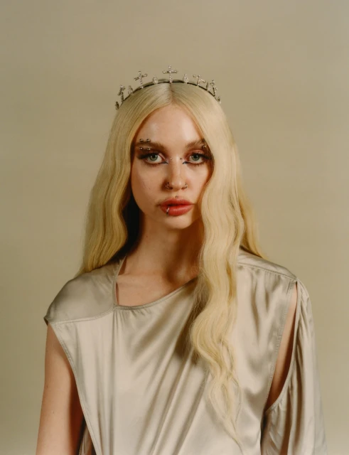 Portrait of a young adult woman with wavy blonde hair wearing a silver crown, a silver dress, and face piercings against a neutral backdrop.