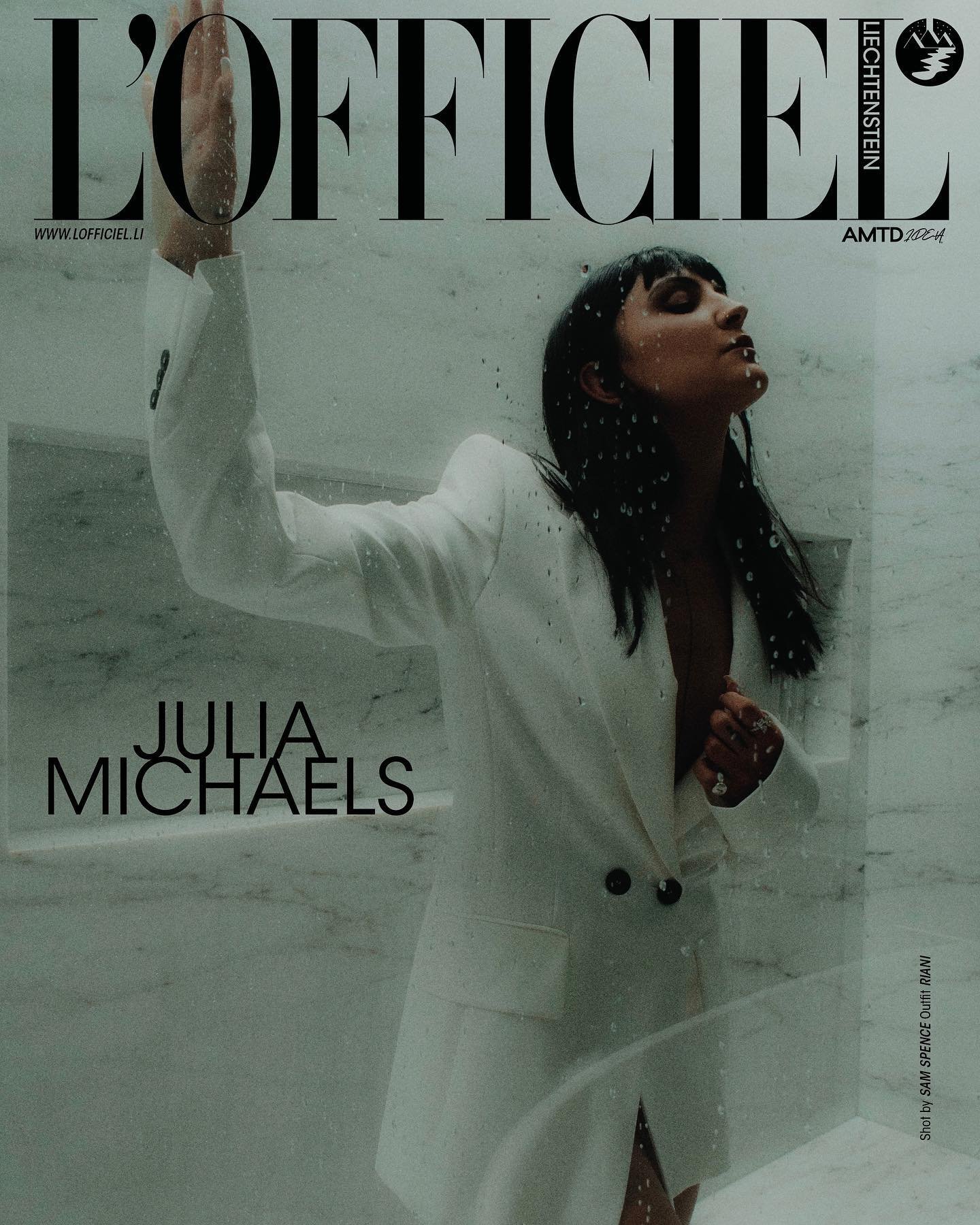 Julia Michaels on L'Officiel Liechtenstein cover in a white suit. Portrait features an eye-level angle, and a marble-like background.
