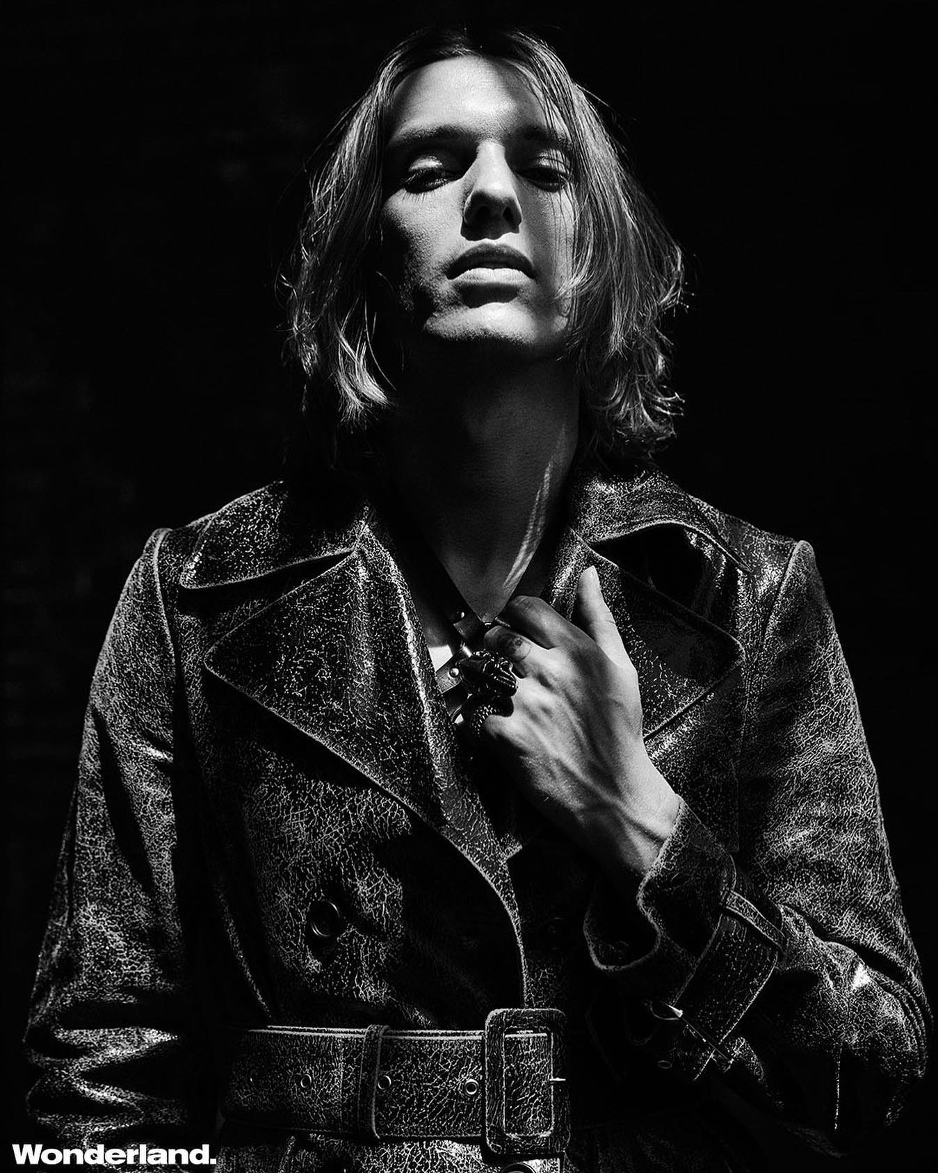 Black and white portrait of a fair-skinned adult male with long hair wearing a textured coat and ornate ring against a dark background.