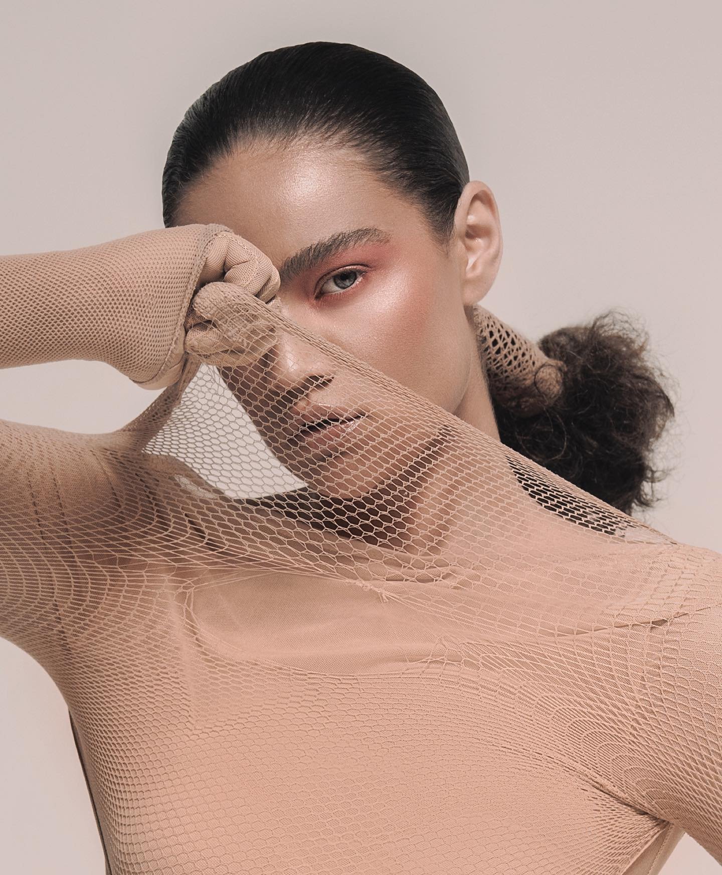Close-up studio portrait of a woman with pink makeup and a tan mesh bodysuit holding the mesh material over part of her face.