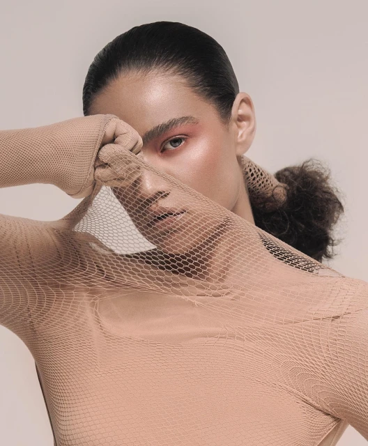 Close-up studio portrait of a woman with pink makeup and a tan mesh bodysuit holding the mesh material over part of her face.
