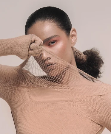 Close-up studio portrait of a woman with pink makeup and a tan mesh bodysuit holding the mesh material over part of her face.