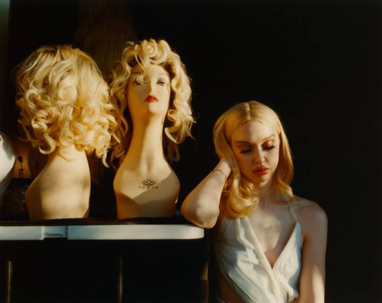 Woman with blonde hair and red lipstick stands next to mannequin heads with blonde wigs under warm light against a dark backdrop.