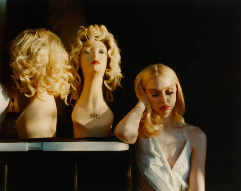 Woman with blonde hair and red lipstick stands next to mannequin heads with blonde wigs under warm light against a dark backdrop.
