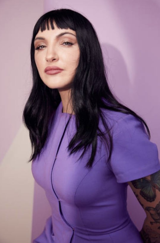 Portrait of a woman with dark hair and bangs wearing a purple dress against a lavender background in a photo studio setting.