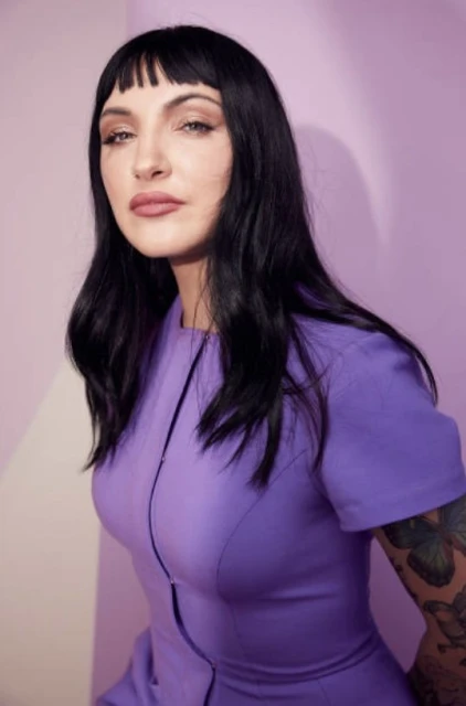 Portrait of a woman with dark hair and bangs wearing a purple dress against a lavender background in a photo studio setting.