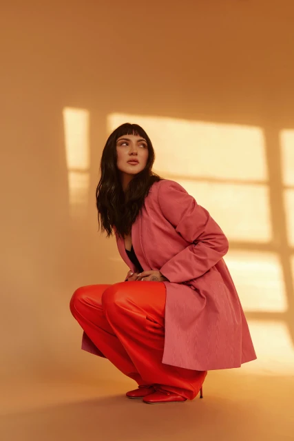 Brunette woman with bangs squats indoors in a pink coat and red pants, looking up and to the side with soft window light.