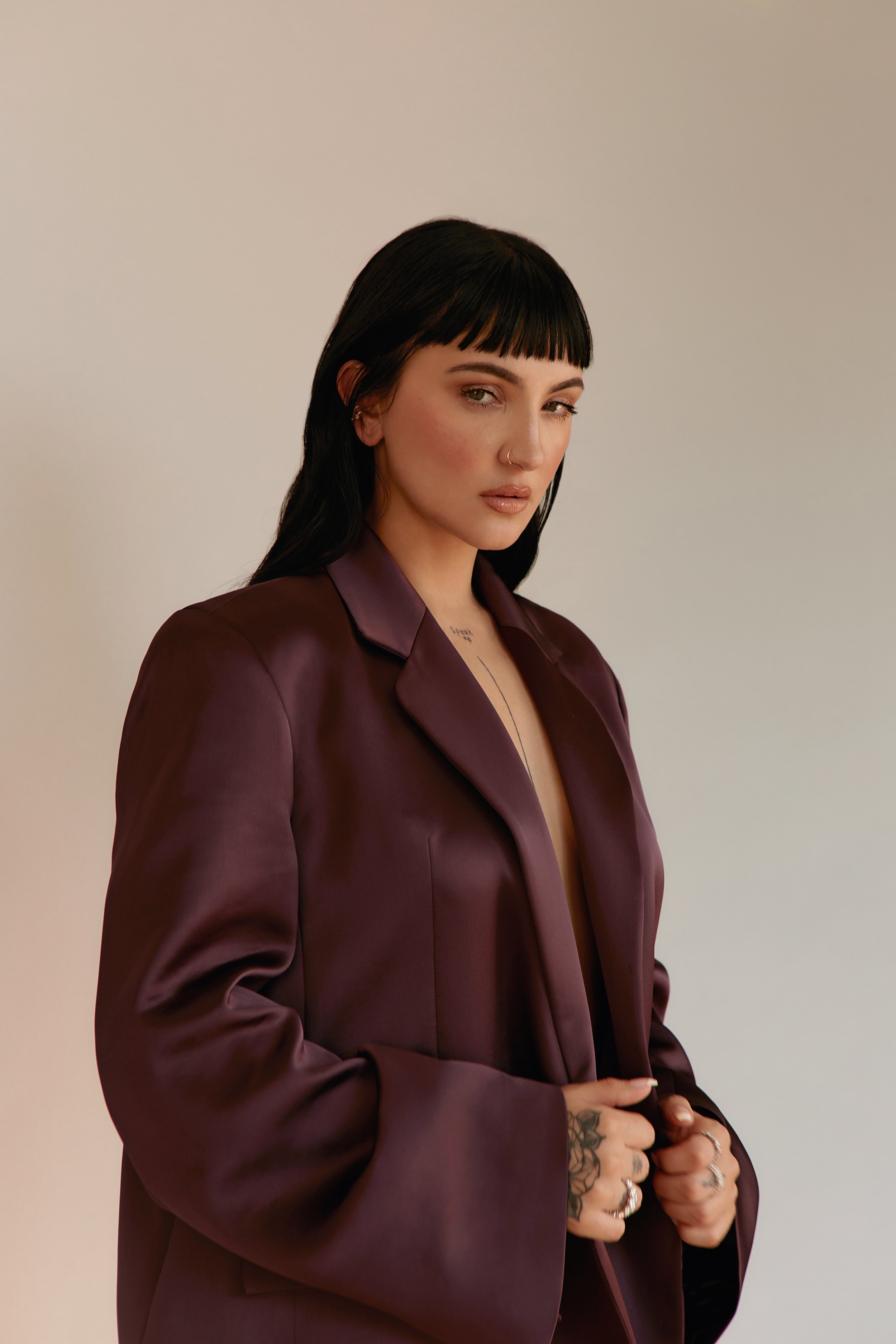 Portrait of a brunette woman with blunt bangs, wearing a plum-colored blazer, posing in a studio with fair lighting and neutral backdrop.
