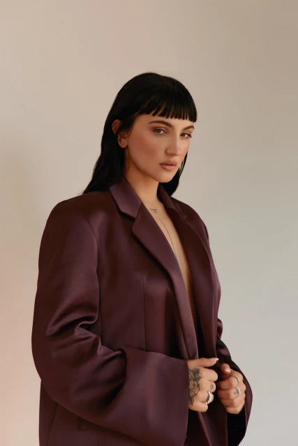 Portrait of a brunette woman with blunt bangs, wearing a plum-colored blazer, posing in a studio with fair lighting and neutral backdrop.