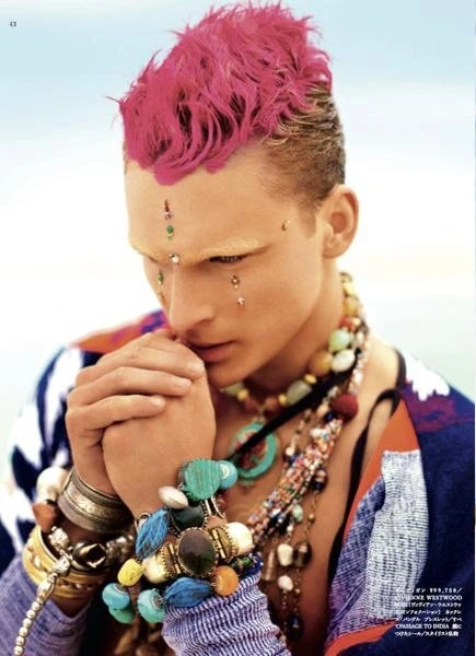 Close-up of a man with pink hair wearing jewelry, bracelets, and an ornate top with a sky background, fashion editorial.