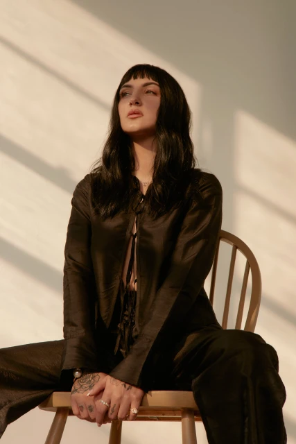 Portrait of a dark-haired, light-skinned woman with bangs in a black satin suit, sitting on a wooden chair in a minimally styled setting.