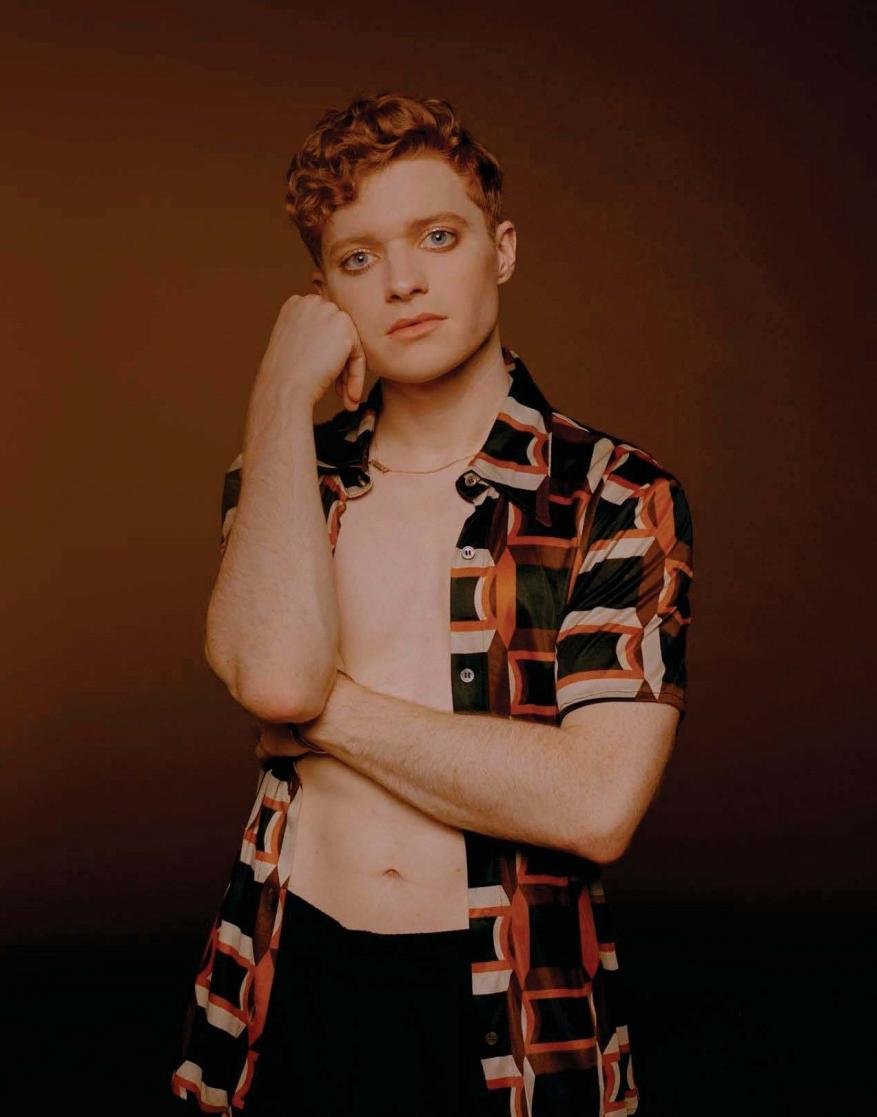 Red-haired person in an open retro-patterned shirt, fair skin, and dark eye makeup posing against a warm brown studio backdrop.