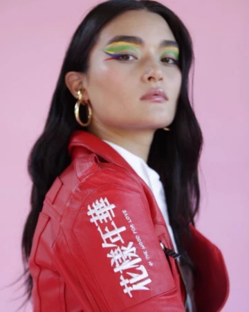 Woman wearing red leather jacket with white Chinese lettering and colourful eye makeup against pink background