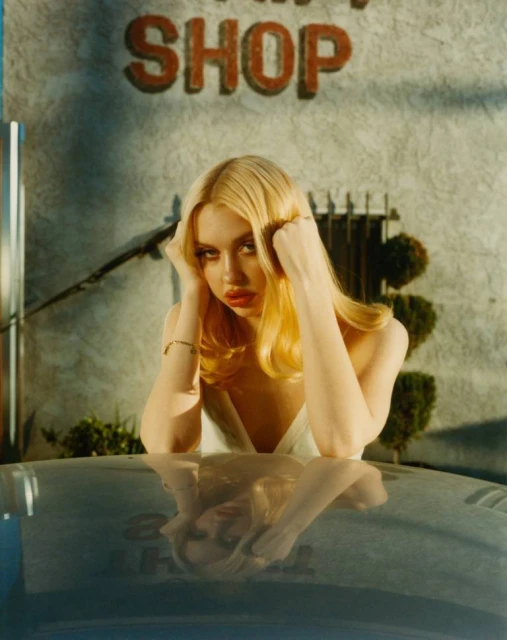 Blonde woman with bright red lips rests her head on her hands, leaning over a car, with a faded building and tree in the background.