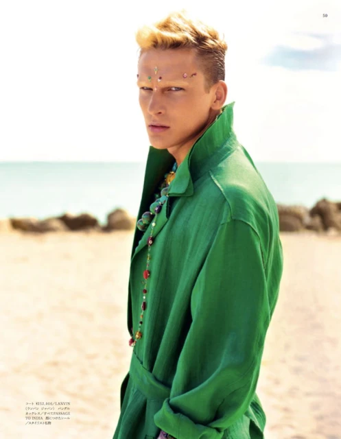 Man with blond hair and jeweled forehead wears a loose green shirt and beaded necklace on a beach with a blue ocean backdrop.