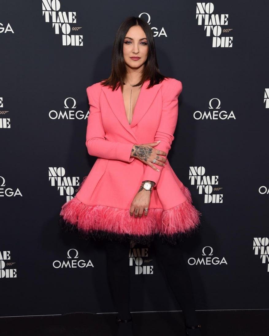 Woman in pink blazer dress poses in front of 'No Time To Die' backdrop, dark hair, watch, tattoos. Event photo with Omega logos.