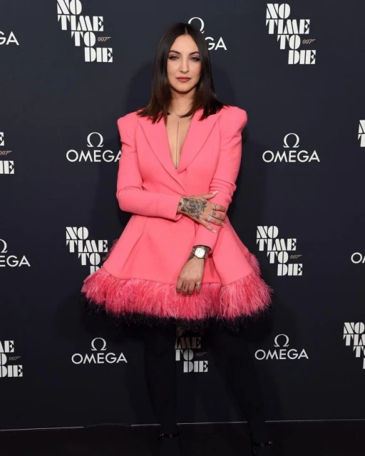Woman in pink blazer dress poses in front of 'No Time To Die' backdrop, dark hair, watch, tattoos. Event photo with Omega logos.