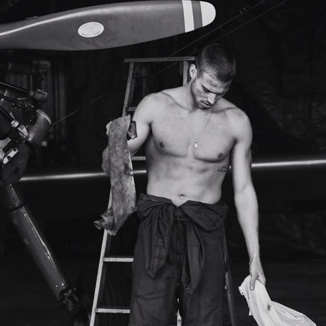 Monochrome portrait of a shirtless man in coveralls holding rags in an airplane hangar near an airplane and ladder.