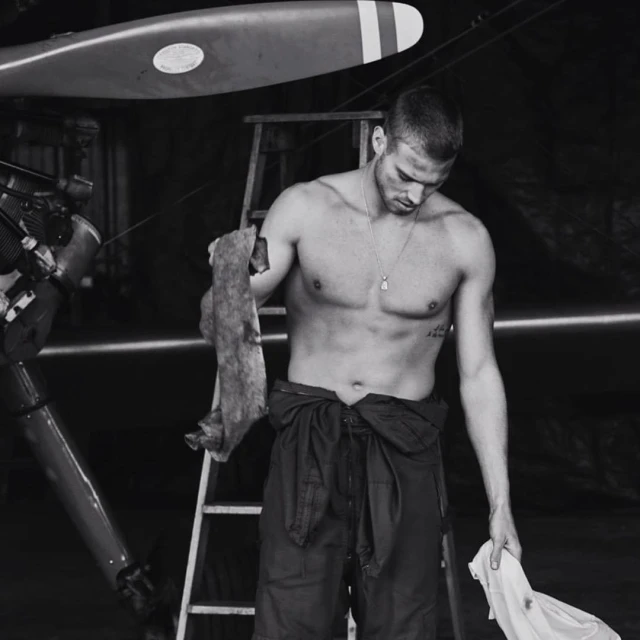 Monochrome portrait of a shirtless man in coveralls holding rags in an airplane hangar near an airplane and ladder.