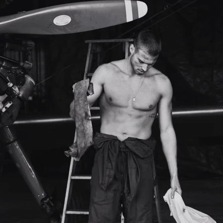 Monochrome portrait of a shirtless man in coveralls holding rags in an airplane hangar near an airplane and ladder.