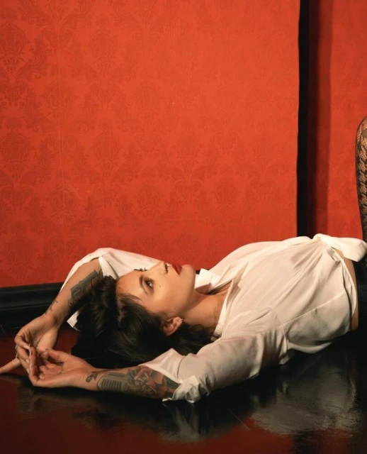 Adult woman with tattoos poses while lying on a glossy floor in a photo studio with a red patterned wall in the background.