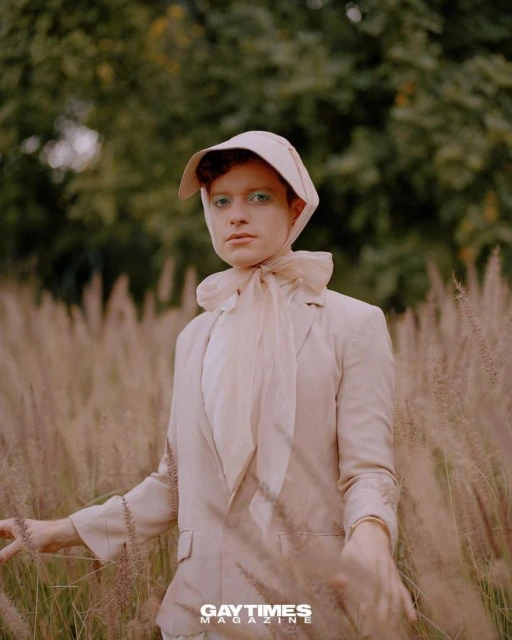 Portrait of a person wearing a cream-colored bonnet, blazer, and scarf while standing in a field of tall, wheat-colored grass outdoors.