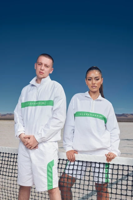 Two people in white and green athletic outfits stand near a tennis net against a blue sky and desert backdrop, creating a sporty, fashion-forward visual.