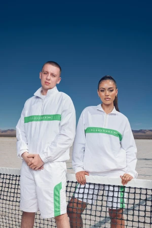 Two people in white and green athletic outfits stand near a tennis net against a blue sky and desert backdrop, creating a sporty, fashion-forward visual.