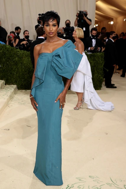 Woman in a teal one-shoulder gown with a large bow posing on a red carpet at an event with people and cameras in the background.