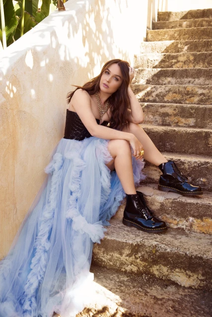 Brunette woman in black top and blue tulle dress sits on stone steps in sunlight, wearing black boots and gold jewelry.