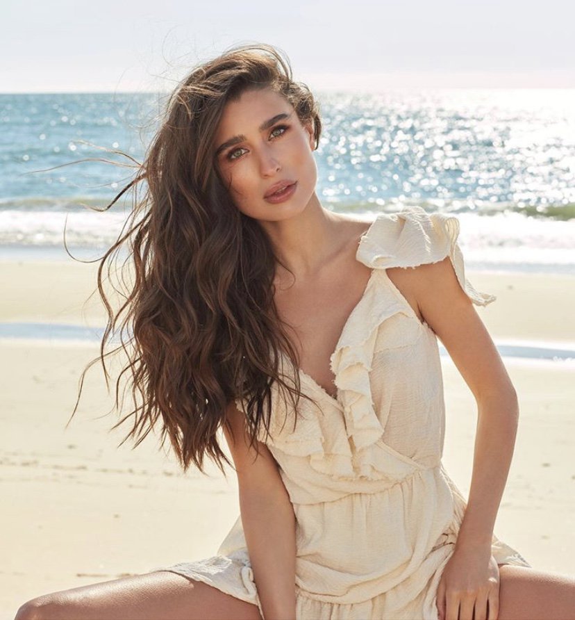 Woman with long wavy brown hair wearing a cream ruffled dress seated on a sandy beach