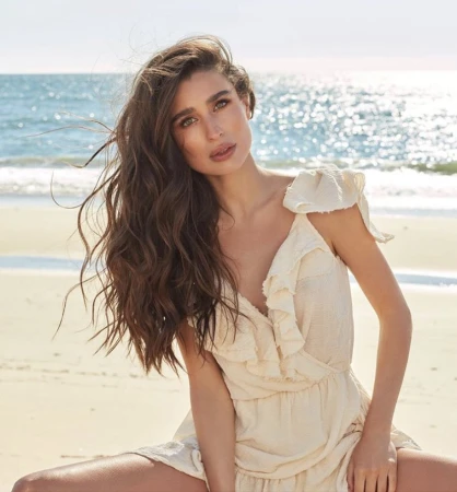 Woman with long wavy brown hair wearing a cream ruffled dress seated on a sandy beach