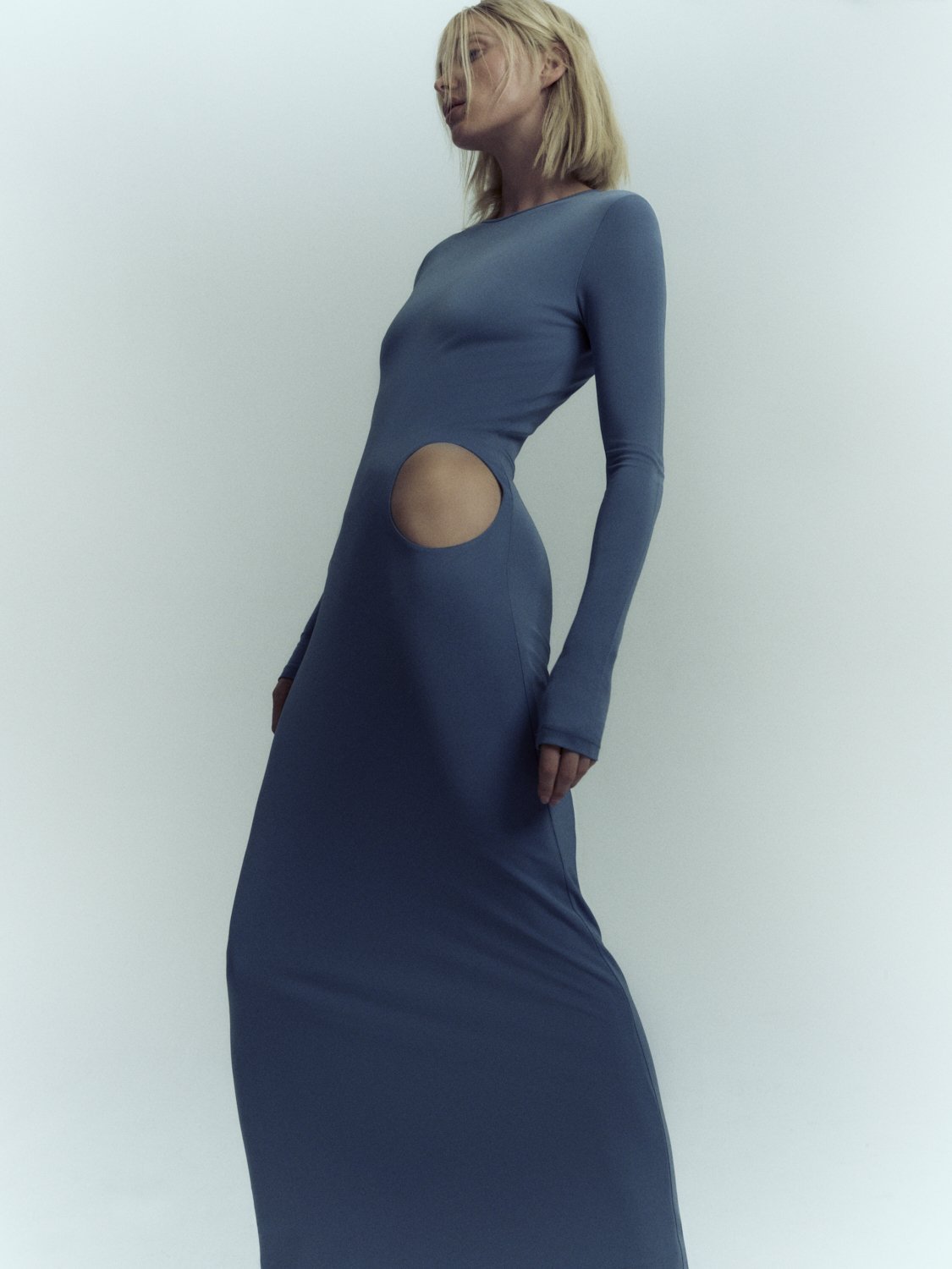 A blonde adult female fashion model poses in a long-sleeved blue dress with a cutout in a studio setting, showcasing contemporary apparel.