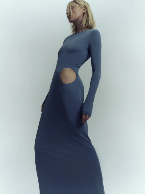 A blonde adult female fashion model poses in a long-sleeved blue dress with a cutout in a studio setting, showcasing contemporary apparel.