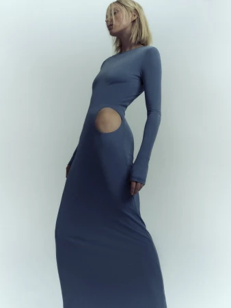 A blonde adult female fashion model poses in a long-sleeved blue dress with a cutout in a studio setting, showcasing contemporary apparel.