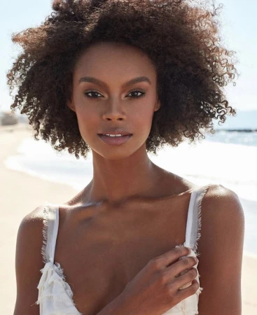 Portrait of a black woman with voluminous curly hair wearing a white textured top against a sunny beach background.
