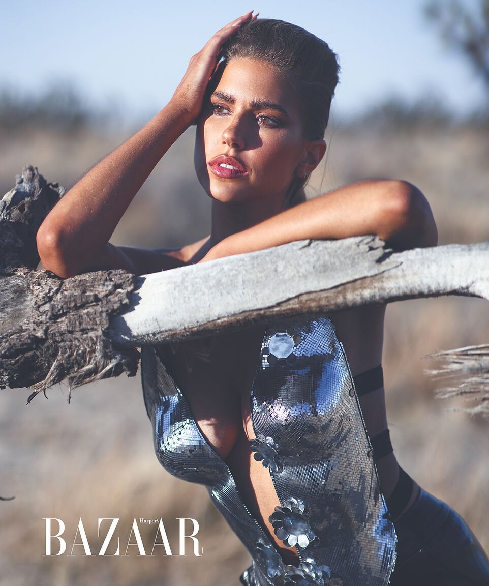A woman in a reflective dress poses in a sunlit outdoor fashion shot with out-of-focus grass and bare branches as a background.