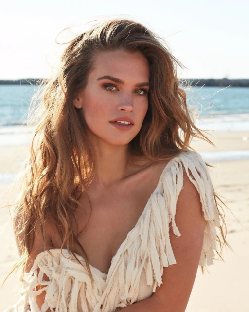 Portrait of a woman on a sunny beach wearing a fringed top. The background includes a calm ocean and a clear sky.