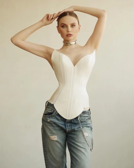 Portrait of a young adult female in a white corset top and ripped blue jeans, silver choker necklace, hands up, in a studio setting.